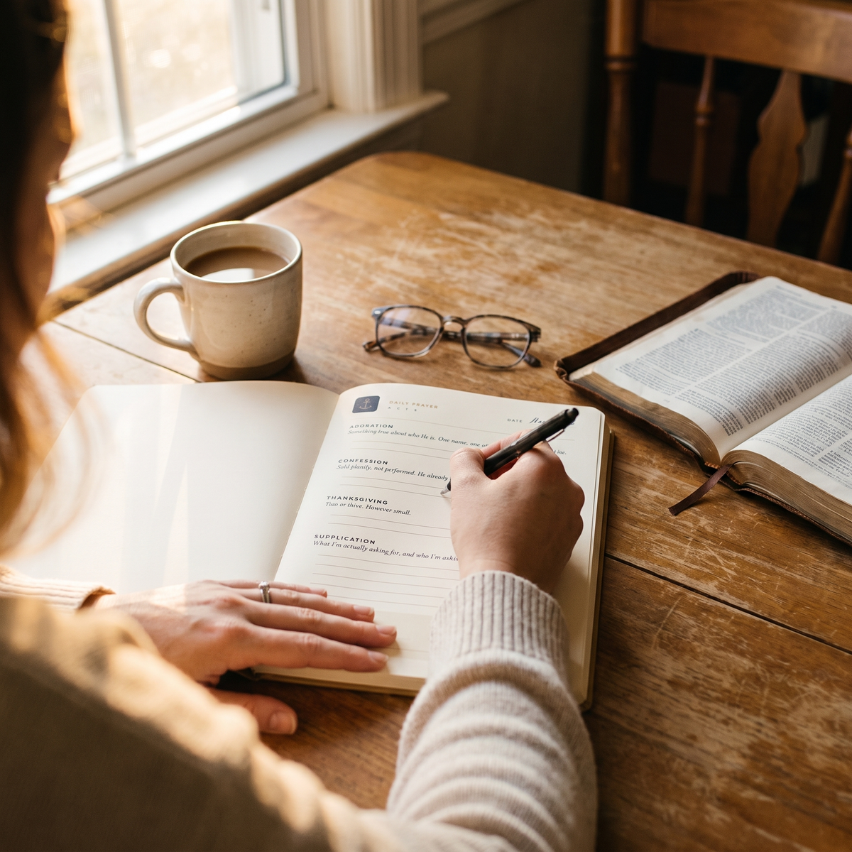 Woman writing in The Anchored Prayer Journal at a kitchen table with coffee and a Bible
